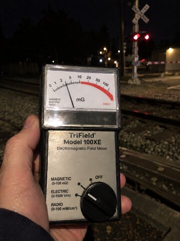 Hand holding TriField 100XE EMF meter near the Washington Street area, reading elevated above ambient background, tracks visible in background